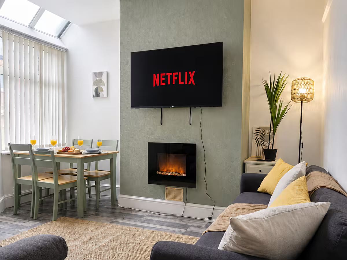 A bright living room: grey sofa with yellow cushions, a wall-mounted television above an electric fire, and a dining table laid for four by the window