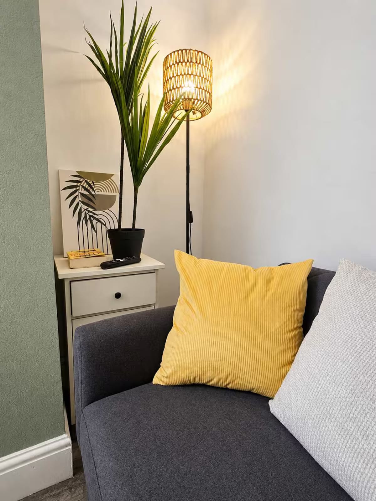 A corner of a sitting room: grey sofa, a yellow cushion, a woven floor lamp and a plant on a side table
