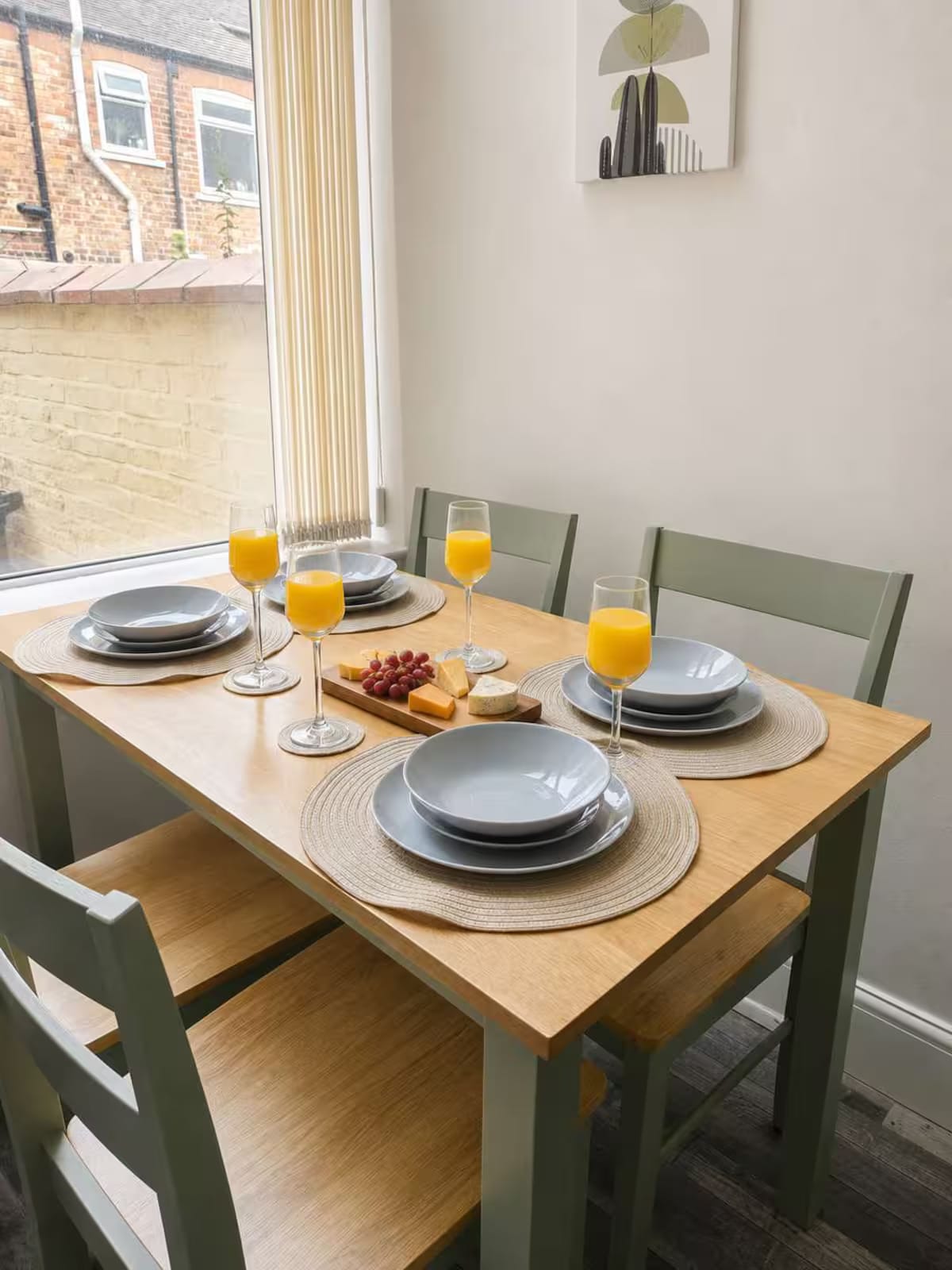 A dining table laid for four with plates, glasses of orange juice and a cheese board, beside a window