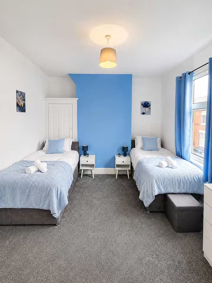 A twin bedroom with two made beds, blue cushions and curtains, and a blue feature wall
